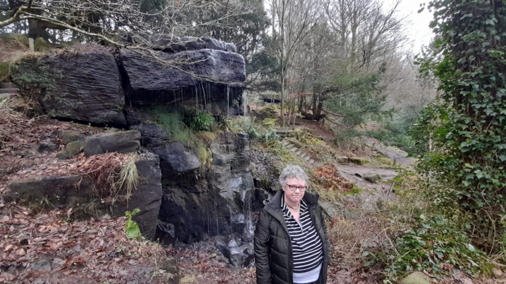 A wide-angle shot of an older woman with glasses and a green padded coat standing in front of a rock formation with small waterfalls. The area is surrounded by woodland with moss-covered rocks, trees, and a forest path leading further into the scene.