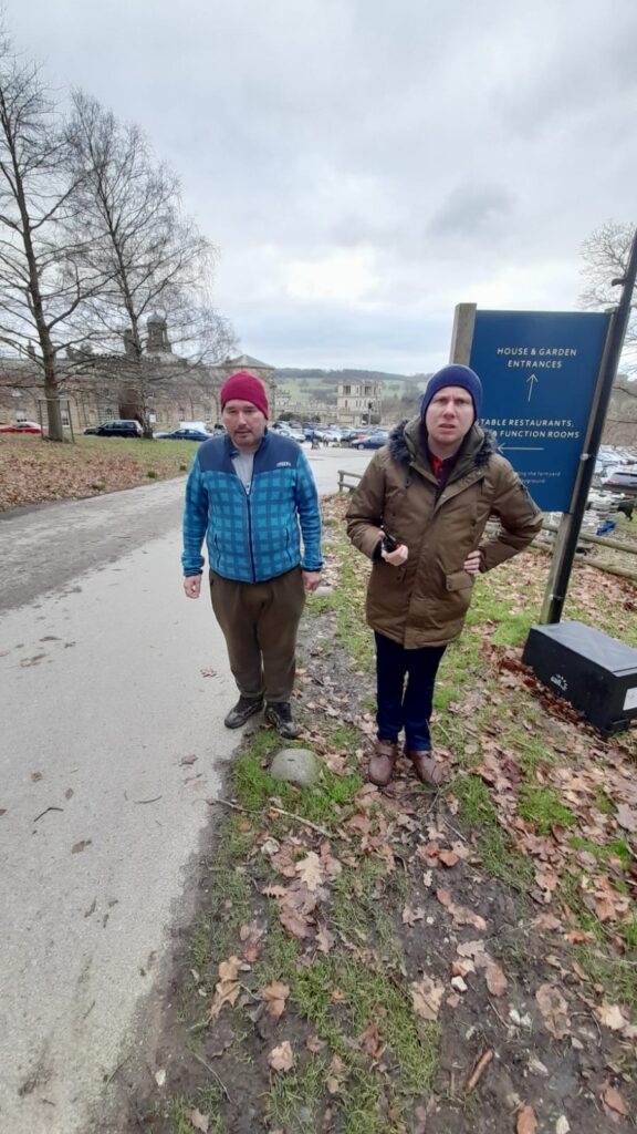 Four people pose in front of the Chatsworth House grounds. The group includes an older woman in a green coat, a woman in a grey coat and yellow t-shirt, a man in a red beanie and blue checkered jacket, and another man in a brown coat holding a small device. The sign behind them points toward the House & Garden entrances.