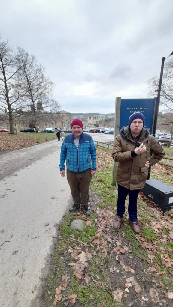 Two men stand on a grassy verge beside a road near Chatsworth House. One man wears a red beanie and a blue checkered jacket, while the other wears a brown coat and navy beanie, holding a small device. A sign reading "House & Garden Entrances" stands nearby, with cars and buildings in the background.