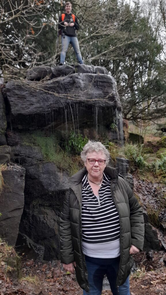 An older woman with glasses and a green padded coat stands in front of a rock formation with small waterfalls. Above her, a person with an orange backpack and jeans stands triumphantly on the rock, smiling. The surrounding area is filled with trees and moss.