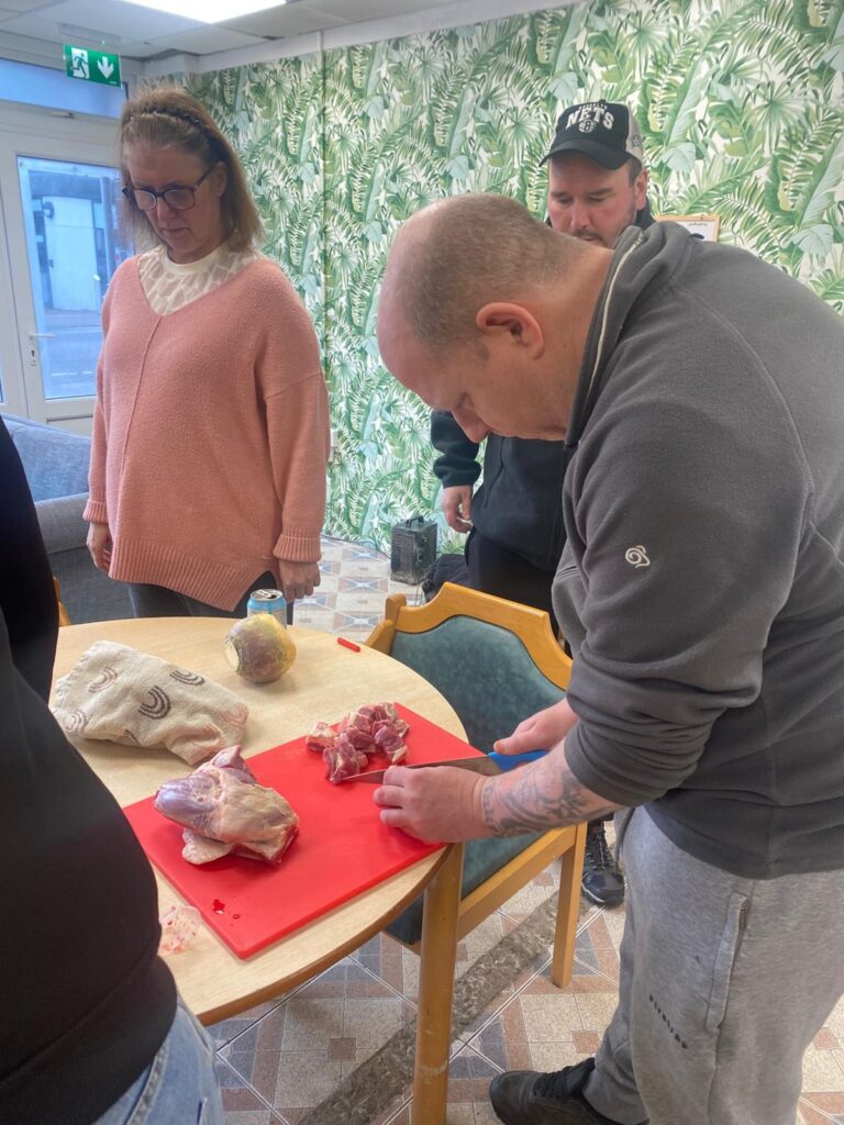 A group scene of a man cutting lamb on a red chopping board, while others, including a woman in a pink jumper, observe and assist in the cooking session.