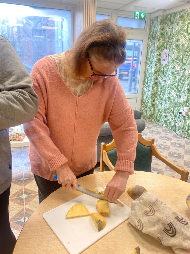 A woman in a pink jumper with glasses chopping swede on a white cutting board, concentrating on preparing the vegetables.