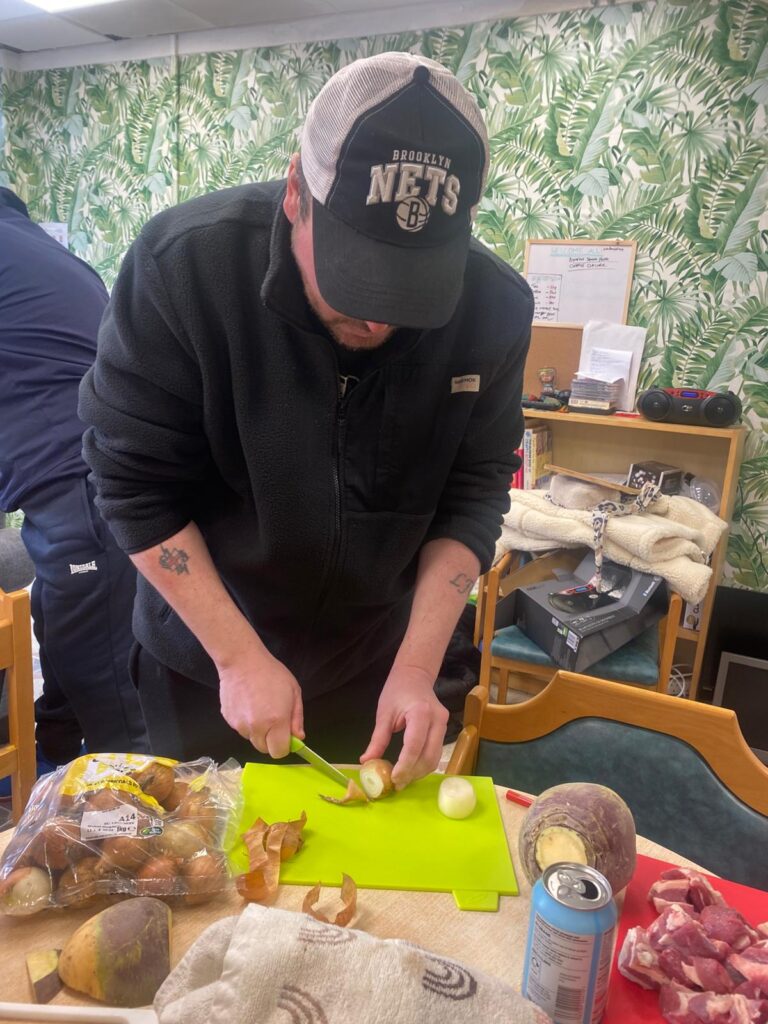 A man in a black cap and fleece peeling an onion on a bright green chopping board, with other vegetables like swede and potatoes nearby.