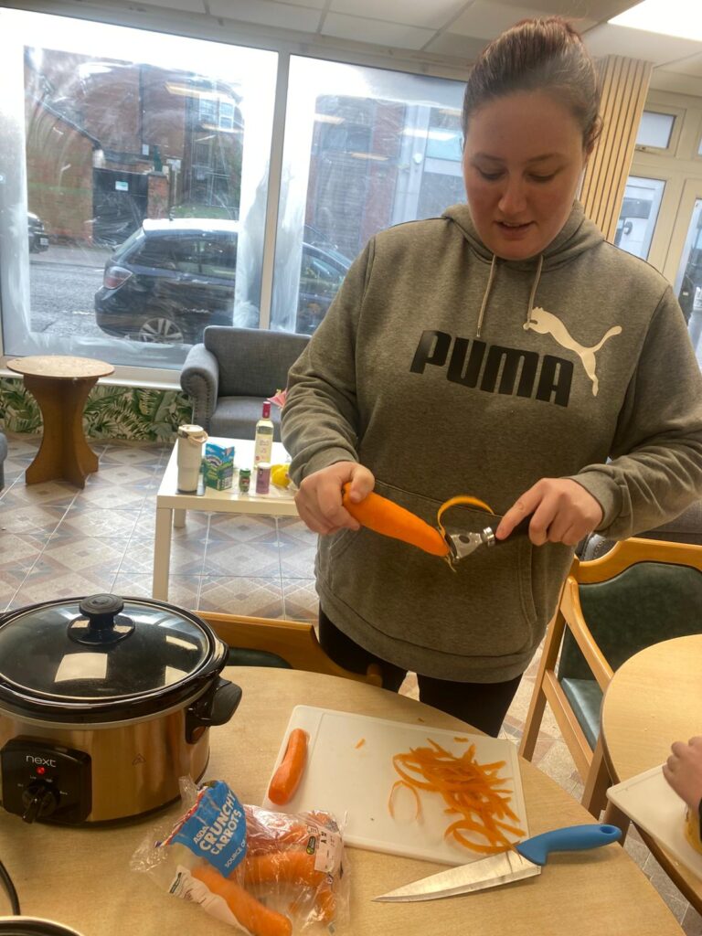 A woman in a grey Puma sweatshirt peeling carrots at a table, with peeled carrot strips and a slow cooker visible on the table.