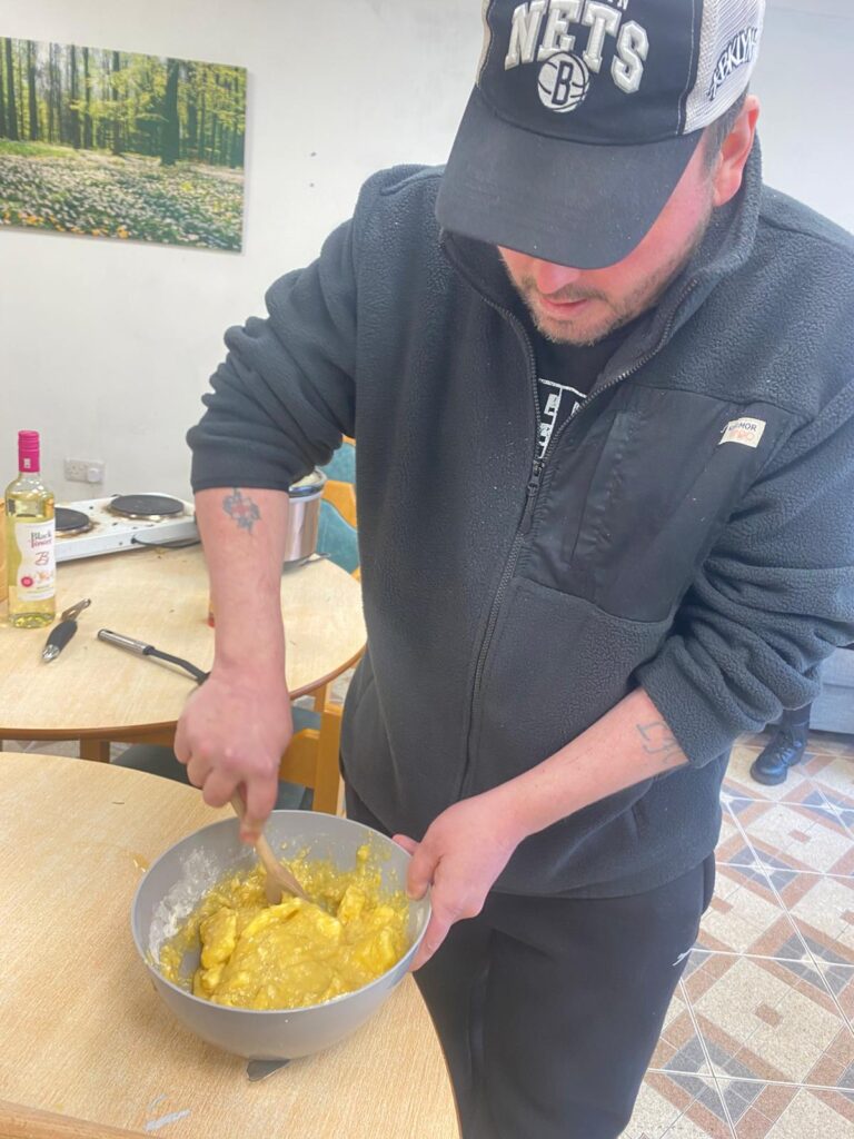 A man in a black Brooklyn Nets cap and fleece stirring a yellow batter mixture in a grey bowl during a cooking session.