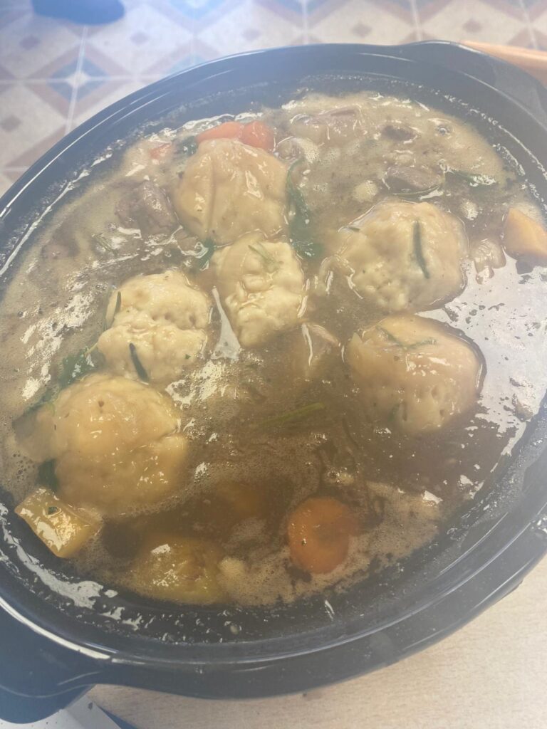 A steaming pot of lamb stew with large, golden dumplings floating on top, surrounded by chunks of carrots and vegetables in a rich broth.