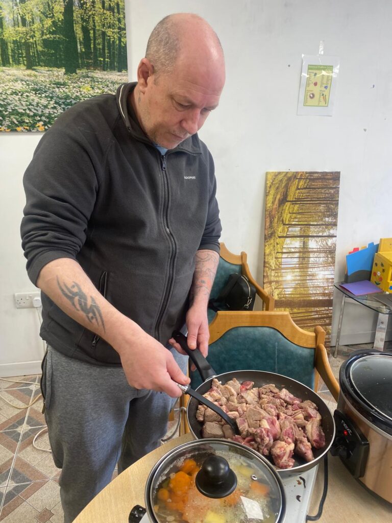 A man in a dark jumper standing at a table, cooking lamb pieces in a frying pan, with a pot of simmering vegetables in the foreground.