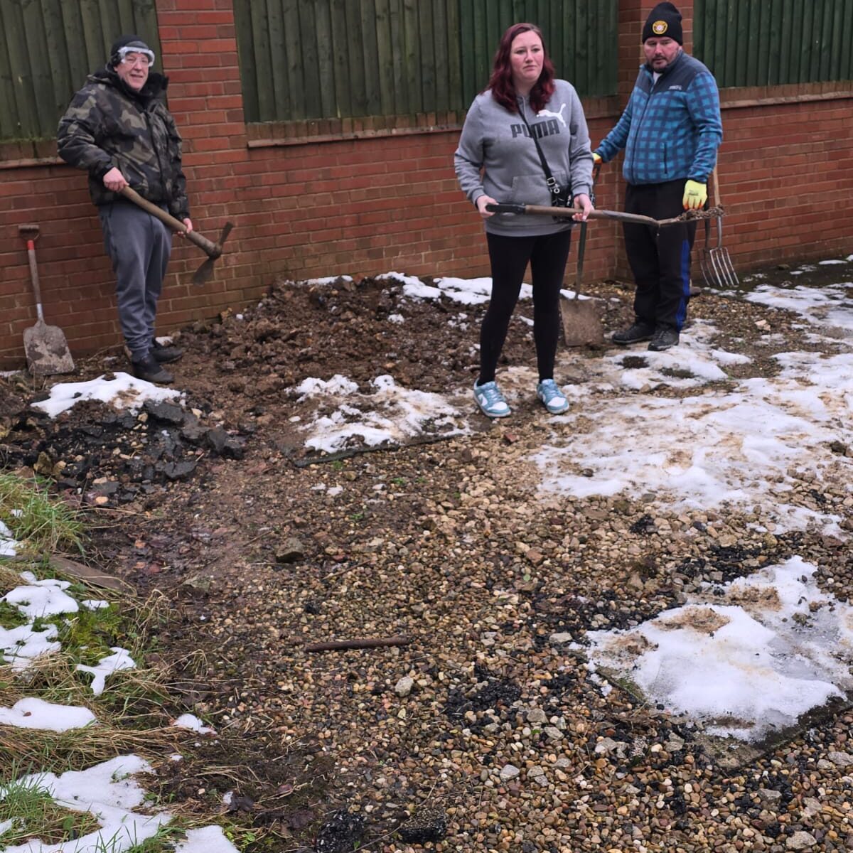 Three individuals working outdoors in a garden area during winter. The ground is covered with patches of snow, gravel, and dirt. One person is holding a pickaxe, another is holding a spade, and the third is holding a garden fork. A brick wall and wooden fence form the background.