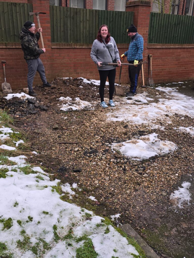 A similar scene showing three individuals in a garden area on a snowy winter day. The ground features patches of snow, gravel, and dirt. The group is equipped with gardening tools, including a pickaxe, spade, and fork, while standing near a brick wall and wooden fence.