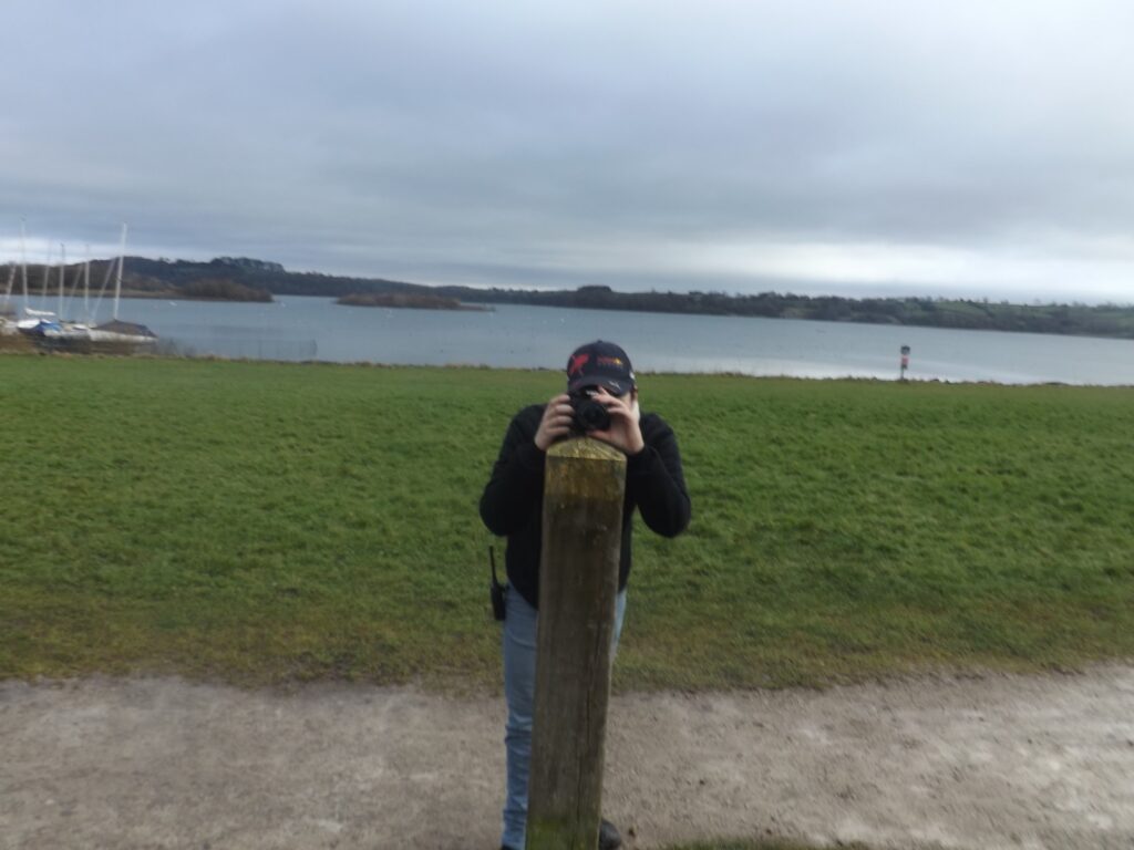 A person standing behind a wooden post, holding a camera, with Carsington Water and sailboats visible in the distance.