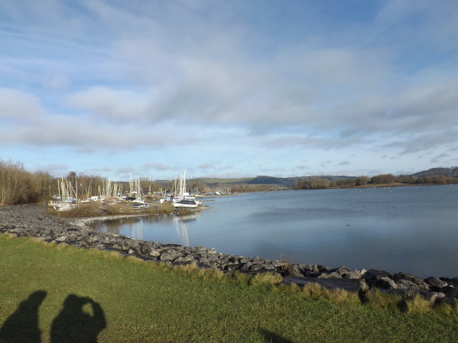 Several sailboats moored along the edge of Carsington Water, surrounded by rocky terrain and grassy fields, with the horizon framed by hills and clouds.