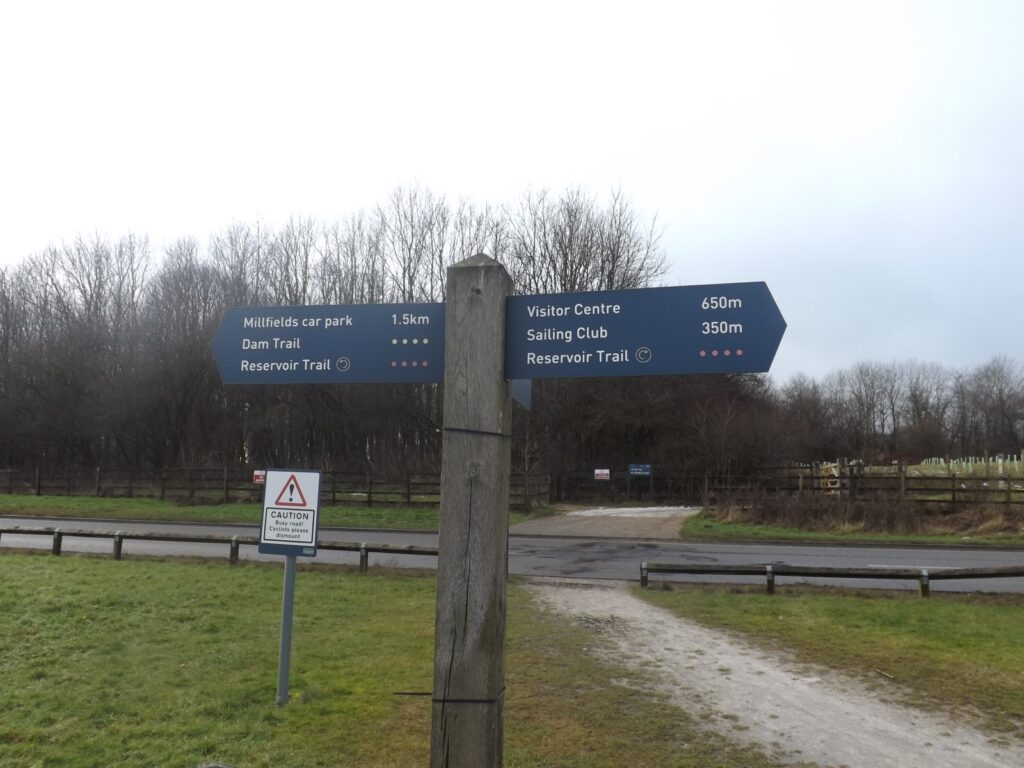 A wooden signpost showing directions to the Millfields car park, Visitor Centre, Dam Trail, and Sailing Club, with trees and a pathway in the background.