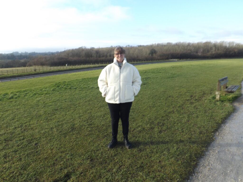 A person wearing a cream-coloured jacket standing on a grassy field with a pathway, wooden bench, and trees in the background, under a bright sky.