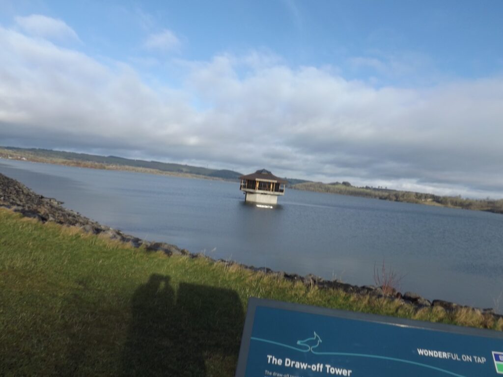 The draw-off tower situated in the reservoir with cloudy skies and surrounding hills, captured from the grassy edge