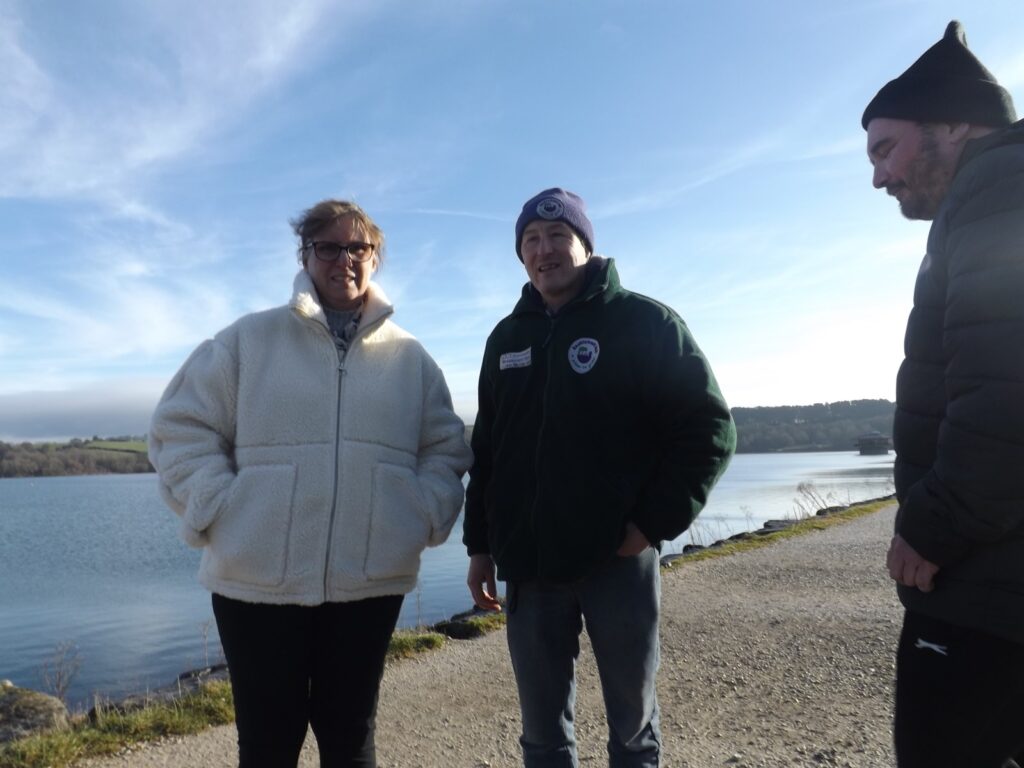 Three people standing on a pathway near the water with a clear blue sky. One is wearing a cream coat, another in a green fleece with a hat, and the third in a dark jacket and beanie, all smiling on a sunny day.