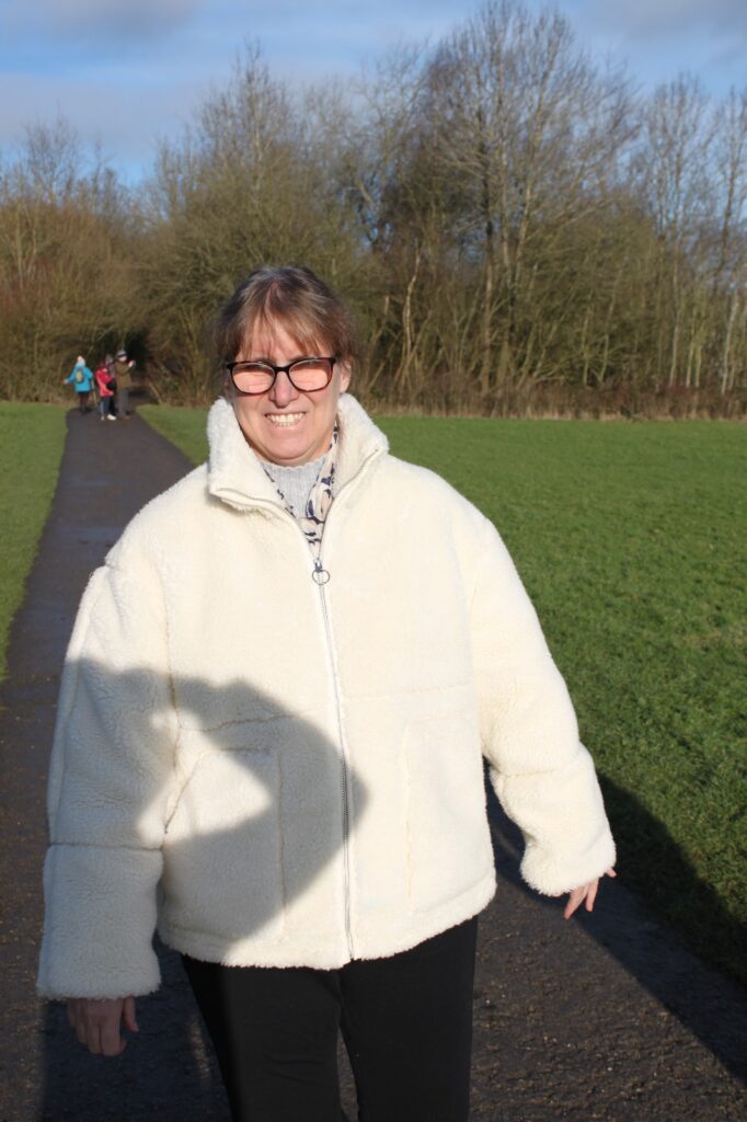 A woman wearing a cream coat and glasses, smiling warmly, standing on a paved path with trees and a grassy field behind her.