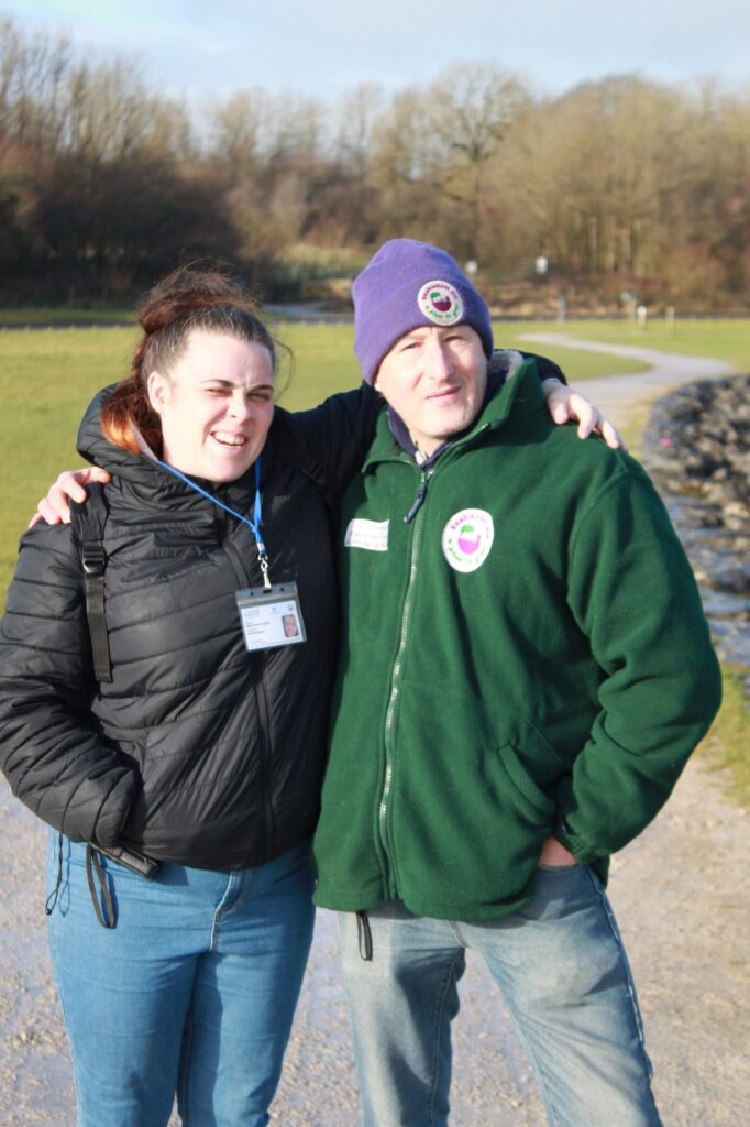 A man in a green fleece and purple hat with his arm around a smiling woman in a black coat, standing on a pathway with trees in the background.