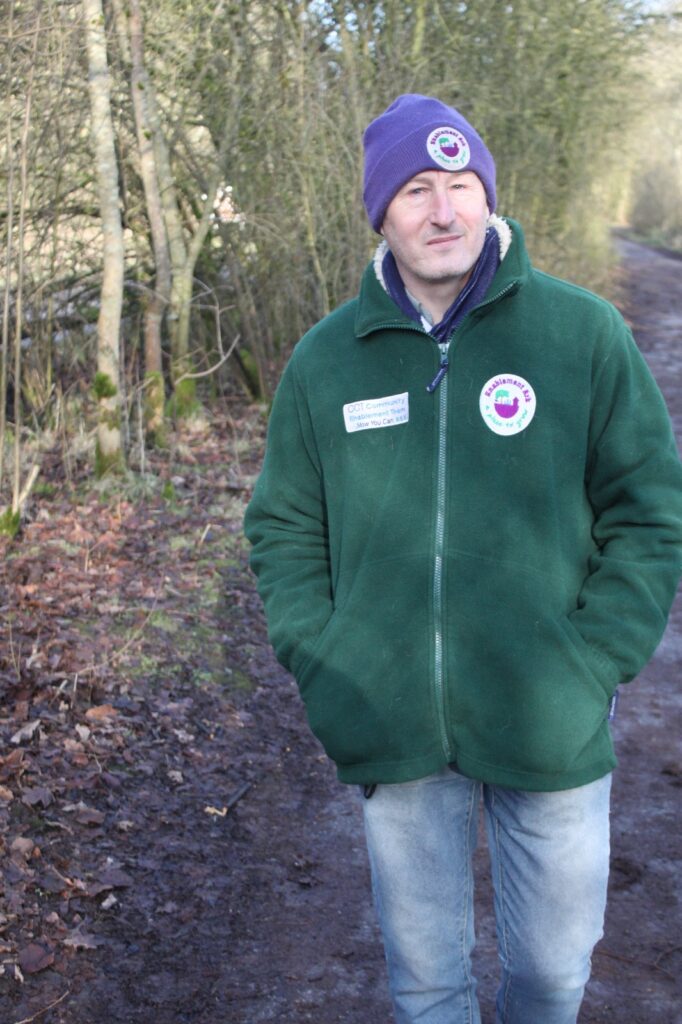 A man in a green fleece and purple hat walking along a muddy forest path with bare trees on either side.