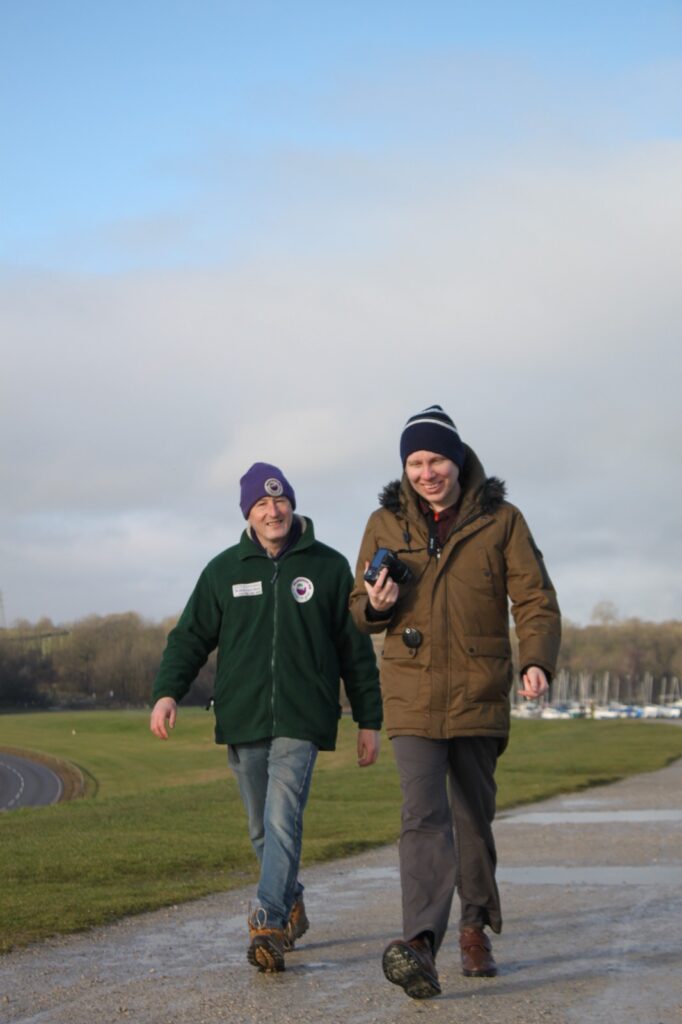 Two men, one in a green fleece and one in a brown coat, walking on a gravel pathway with a grassy field and boats visible in the distance.