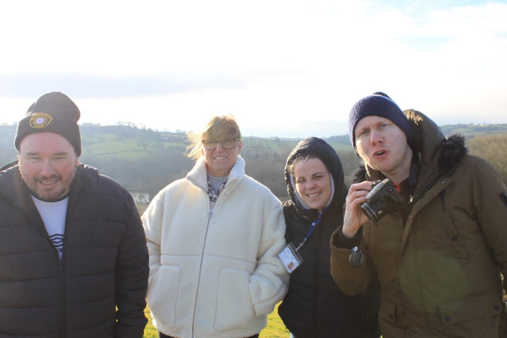 A group of four people in winter coats, smiling and standing close together on a hilltop with a countryside view behind them.