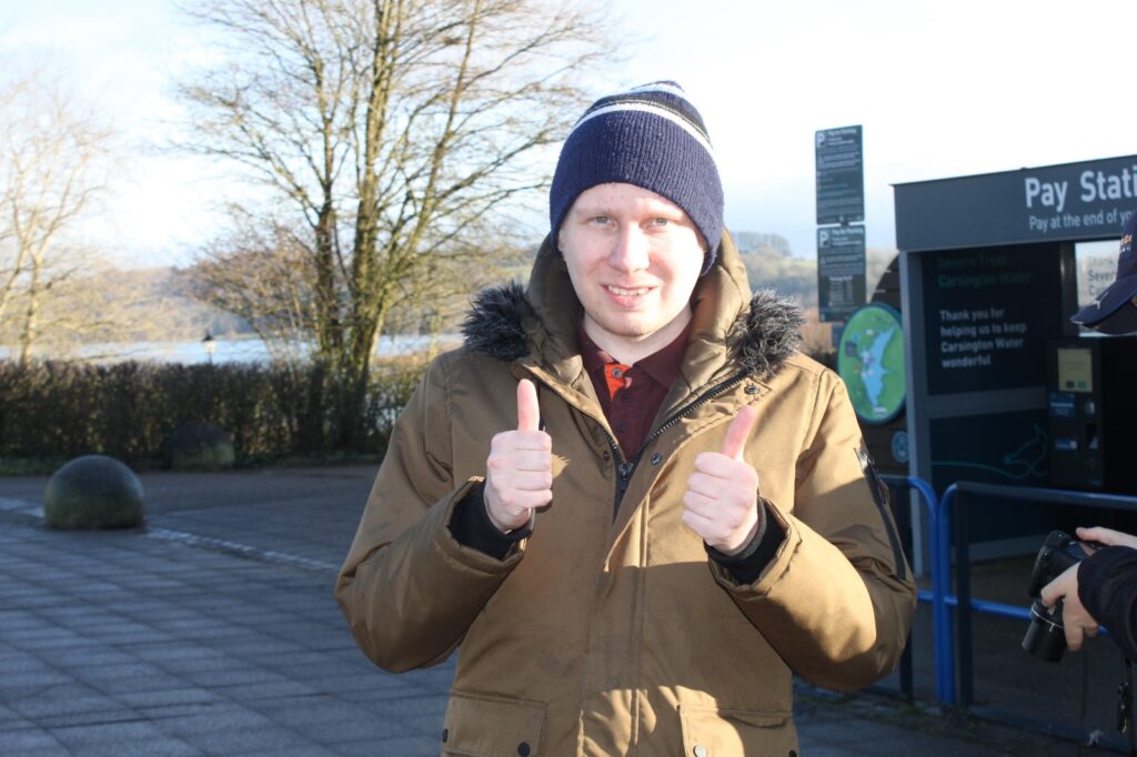 A man in a brown coat and blue hat giving a double thumbs up, standing in front of a pay station and trees with the reservoir in the background.