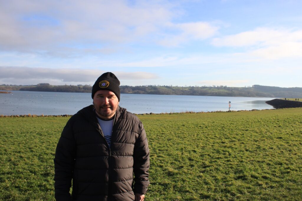 A man in a black coat and hat standing on a grassy field near the edge of the reservoir with hills and a blue sky in the distance.