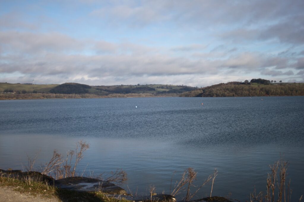 A tranquil view of Carsington Water with still water, grassy hills, and a partly cloudy sky.