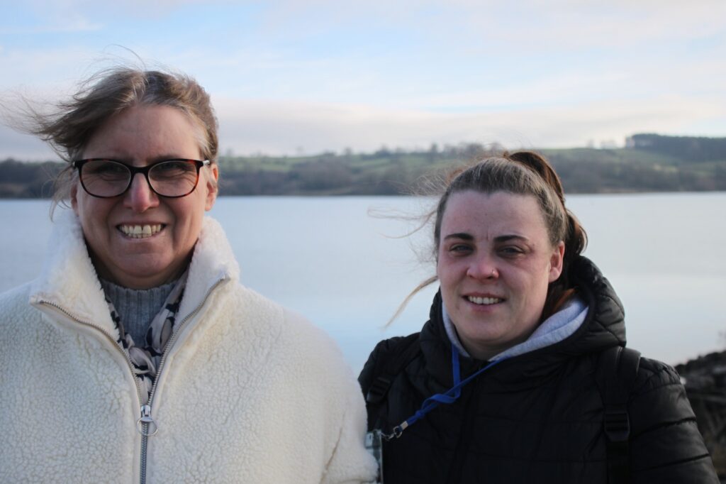 Two women, one in a cream coat and glasses, the other in a black coat, standing close together with the reservoir and hills in the background.