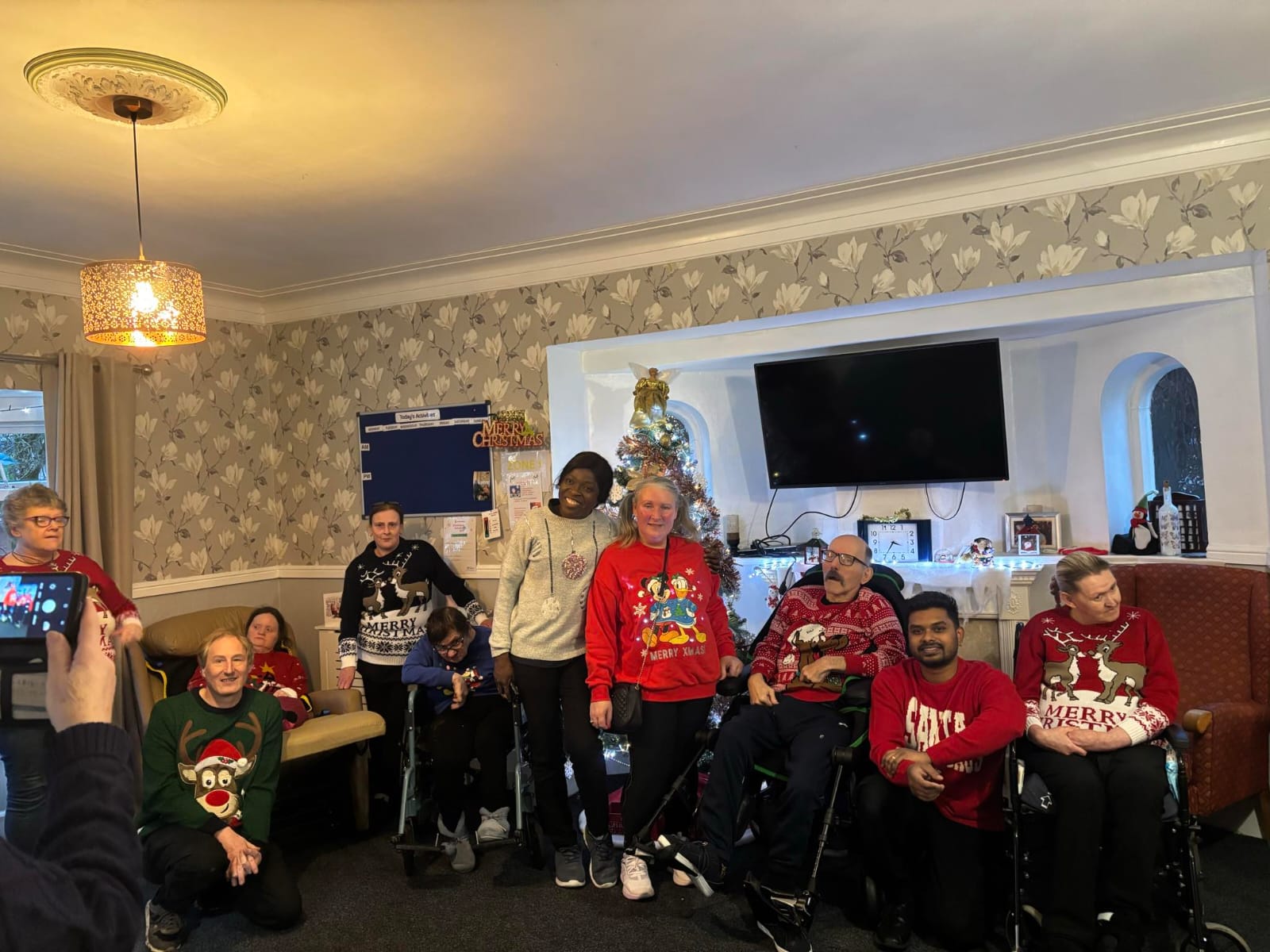 A group of residents and staff from the Community Enablement Team and Enablement Care posing together in festive Christmas jumpers. The group is gathered in a warmly decorated room with a Christmas tree and holiday decor in the background, radiating holiday cheer.