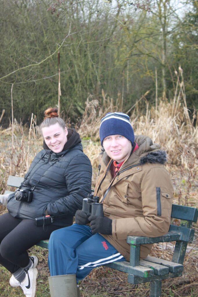 A man and woman sitting on a bench in a natural setting, both smiling at the camera. The man is holding binoculars, and the woman has a camera around her neck.