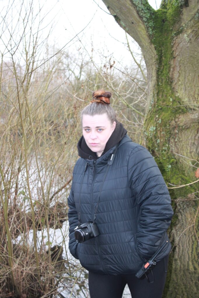 A woman in a black jacket with a camera around her neck, standing near a tree.