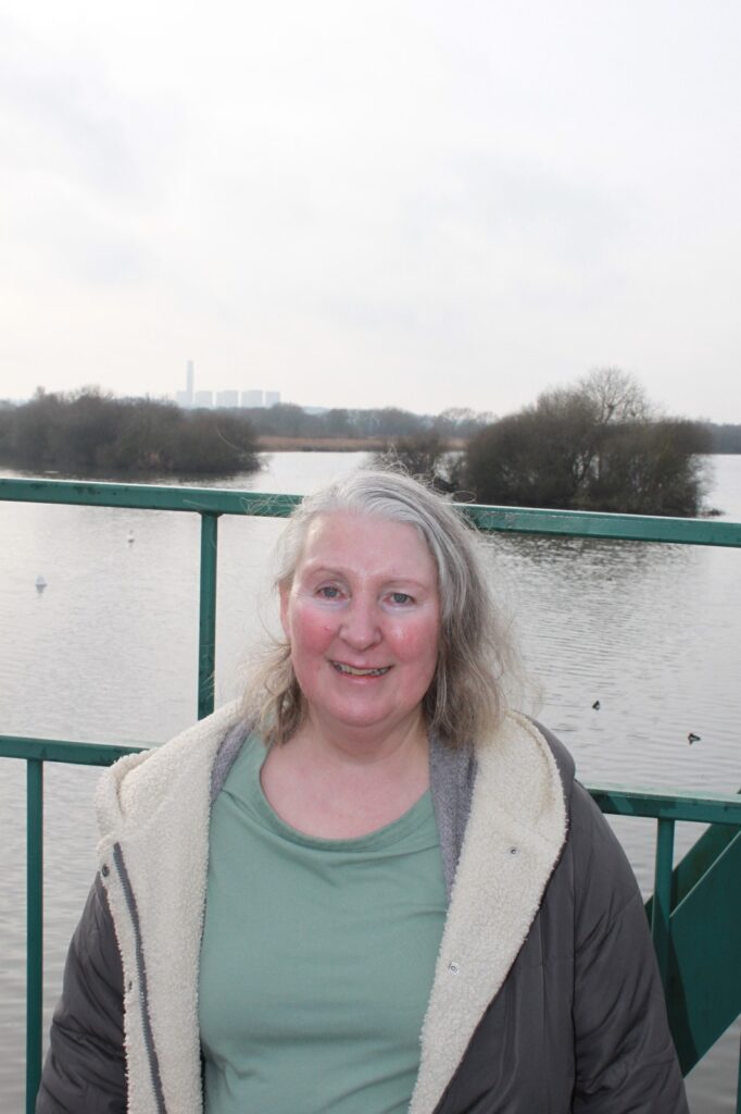 A woman standing on a bridge overlooking a calm river, smiling at the camera with trees and an industrial building visible in the distance.