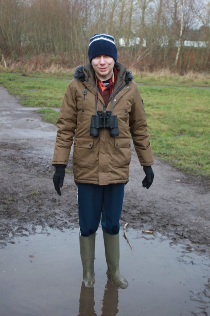 A man wearing wellies and a winter coat standing in a puddle on a muddy path, smiling at the camera with binoculars around his neck.