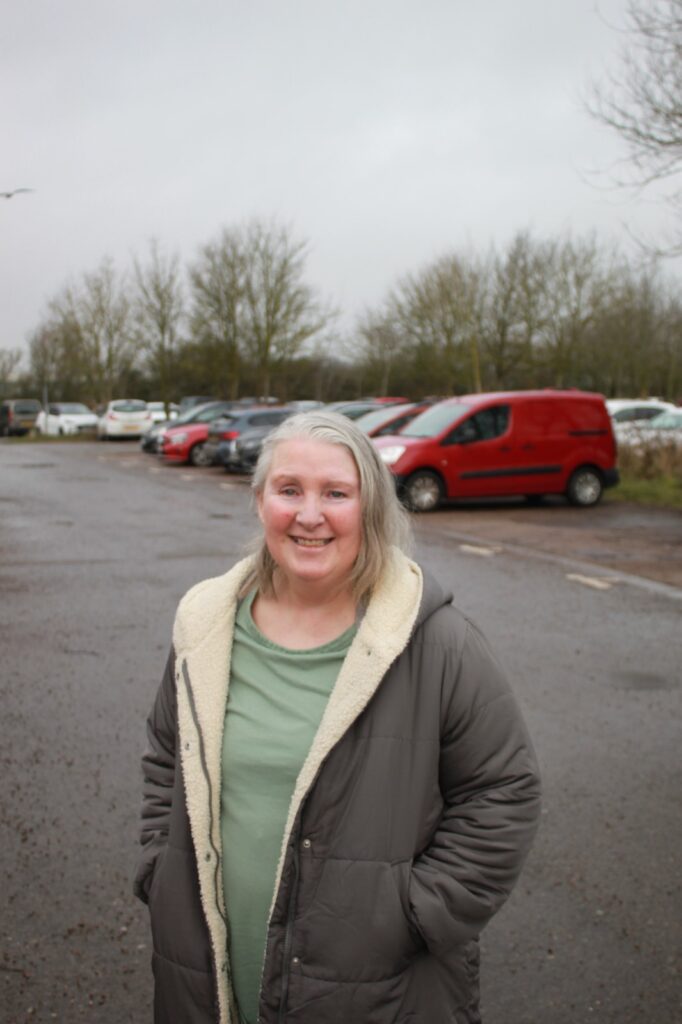 A woman standing in a car park on a cloudy day, smiling warmly at the camera. Rows of parked cars and bare trees are visible in the background.