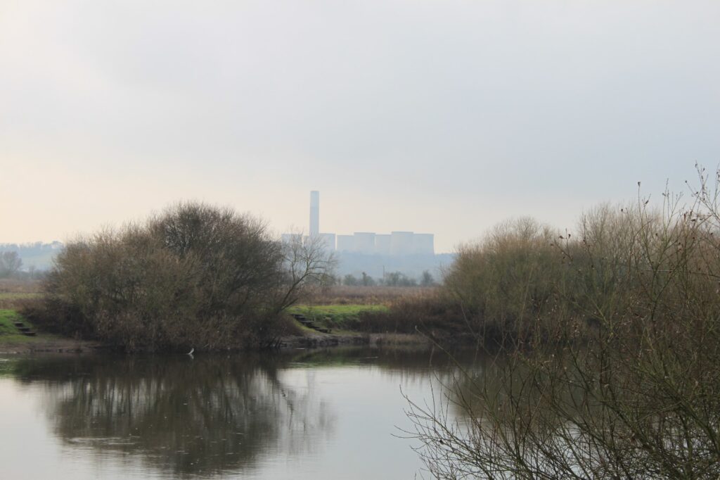 A serene view of a river with trees and a distant industrial building under a cloudy sky.