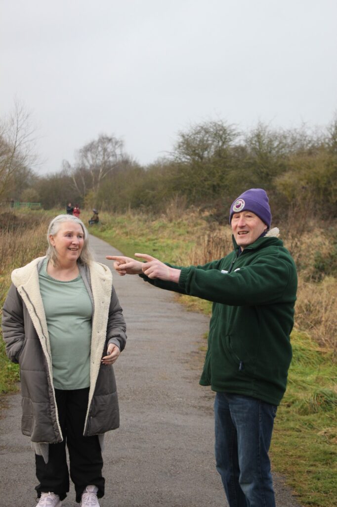 A man pointing something out to a woman on a wooded walking path during the walk.