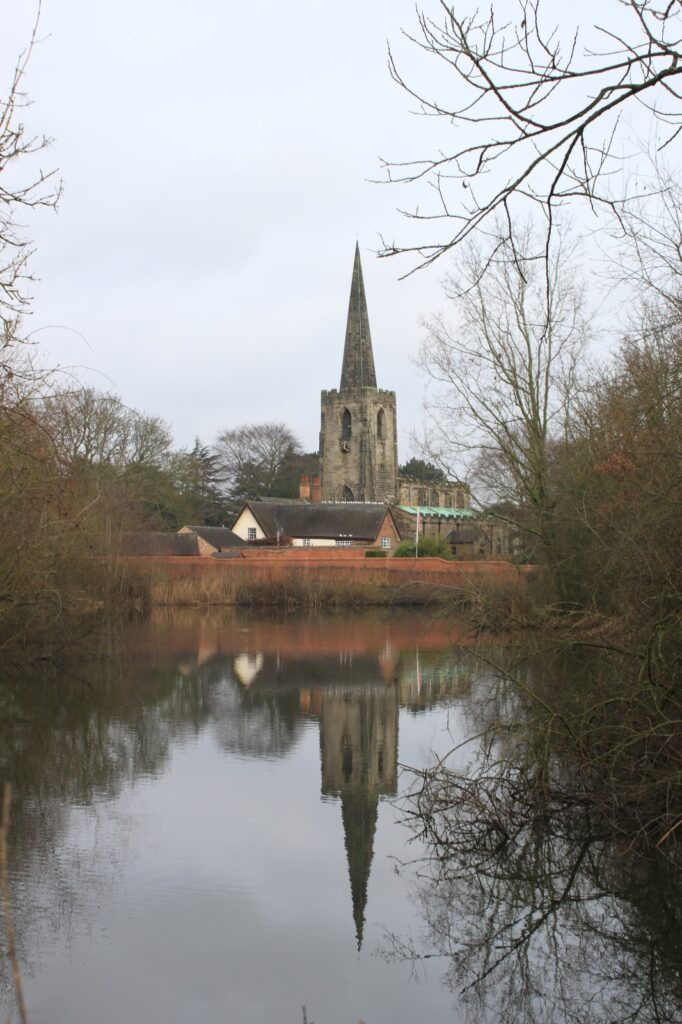 A reflective image of a church steeple, houses, and trees in a calm body of water, framed by winter trees.