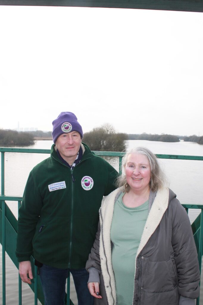 A man and a woman standing on a bridge, smiling at the camera, with a view of the water and trees in the background.