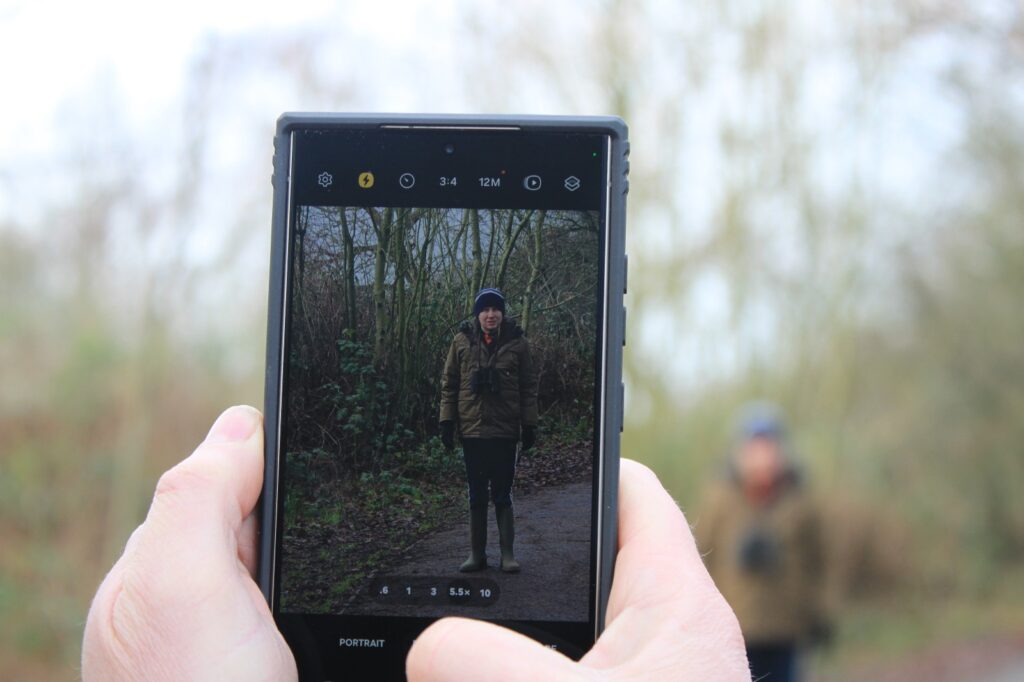 A close-up shot of a phone screen displaying a portrait photo of a man in wellies and a winter coat, with a blurry background of trees.