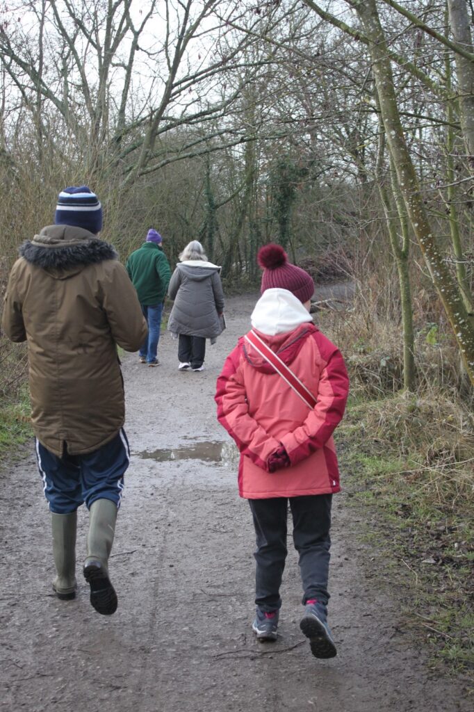 A group of walkers, dressed warmly, strolling along a wooded path on a cloudy winter day.