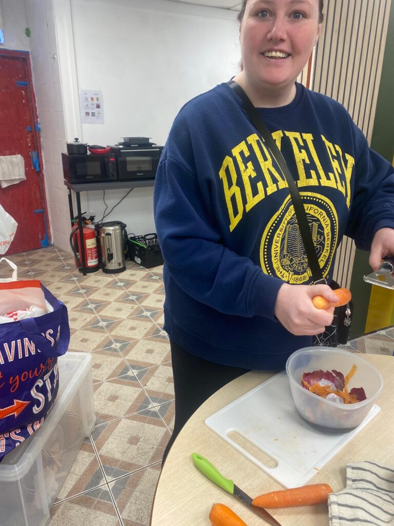 Smiling individual peeling a carrot into a bowl while standing at a table with a chopping board.