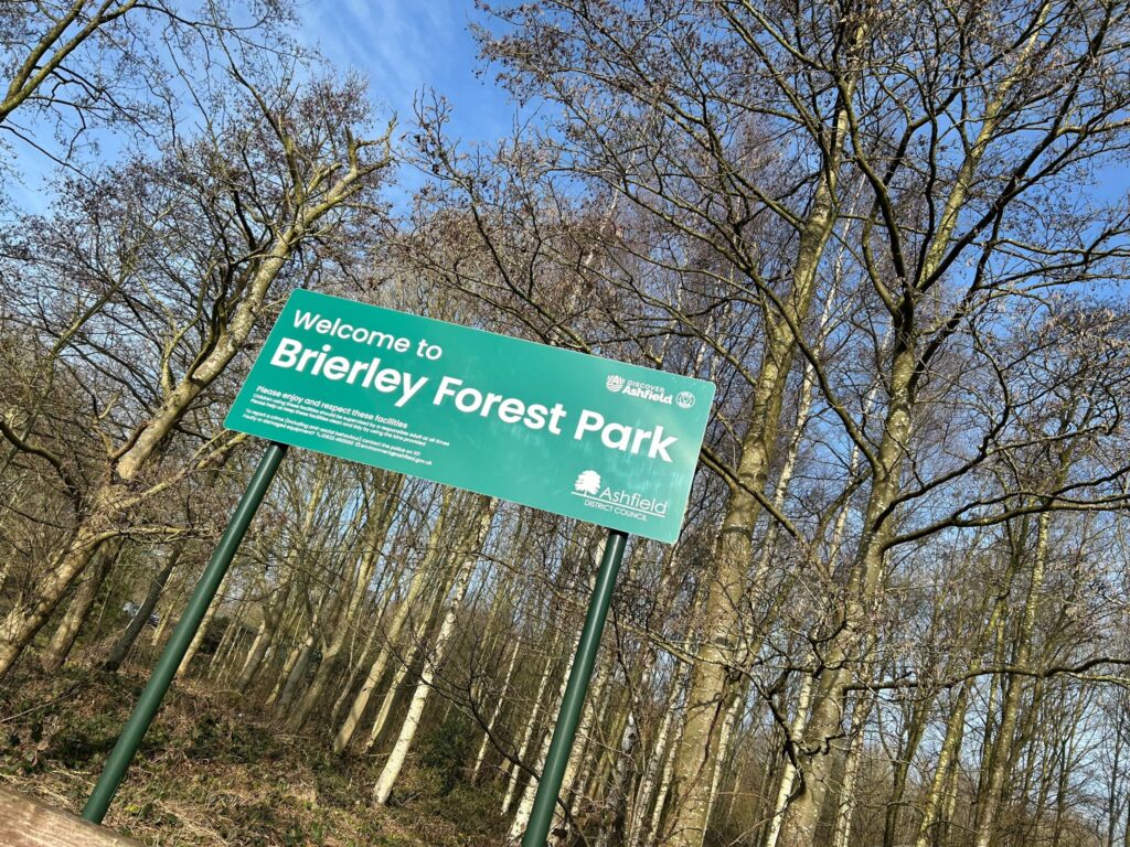 A green sign reading "Welcome to Brierley Forest Park" stands against a backdrop of tall, leafless trees under a clear blue sky.