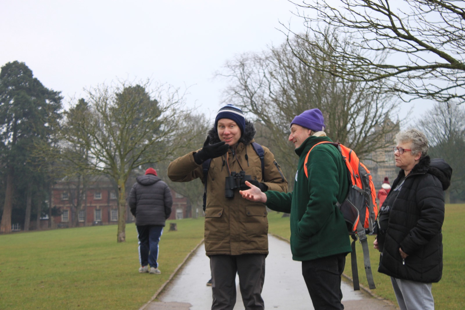 A friendly conversation between two people in warm outdoor gear on a paved path in Wollaton Park, with one gesturing while the other listens. A third person stands nearby observing.