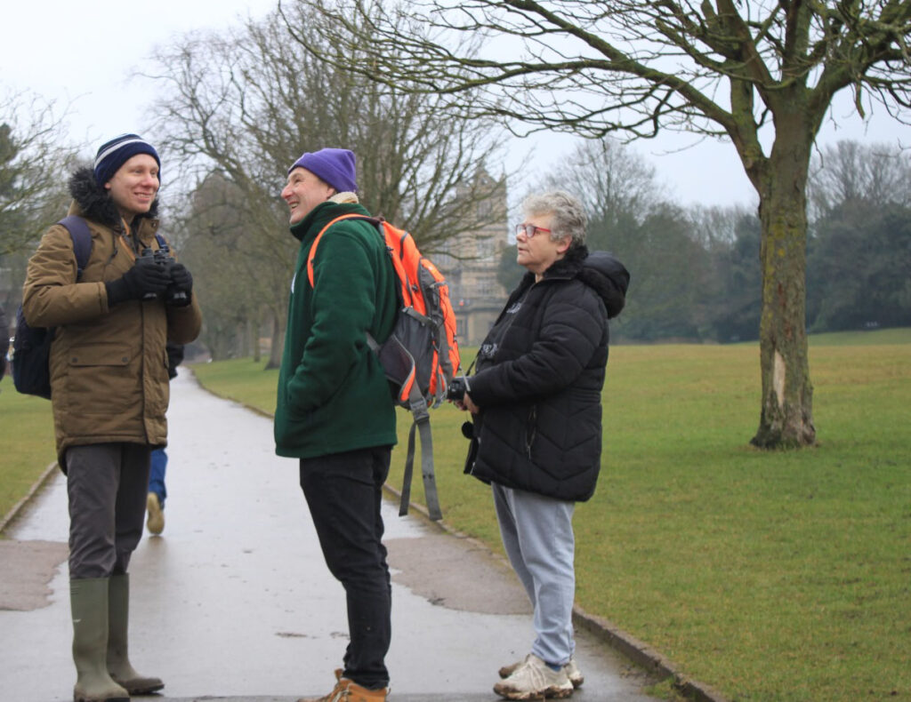 A small group of people engaged in conversation in the park, one person smiling while another listens attentively, with Wollaton Hall visible in the background.