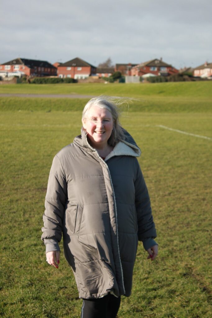 The woman in the grey coat stands on a grassy field, smiling with the wind in her hair. In the background, a row of red-brick houses and a football pitch are visible.