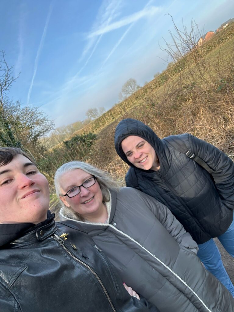 Three people, dressed warmly in jackets and hoodies, pose together for a selfie during their walk along a scenic countryside path, with hedgerows and trees in the background.