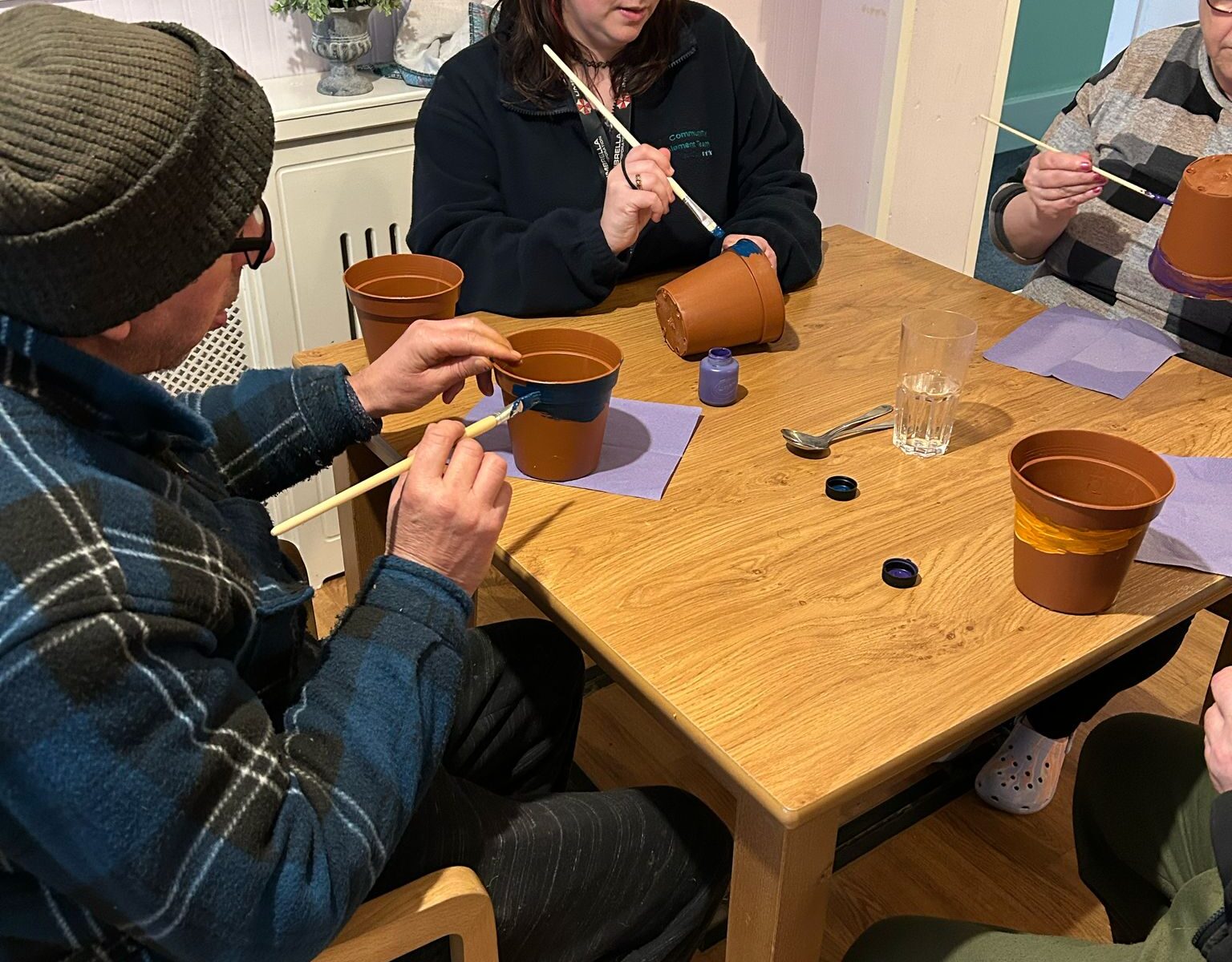 A close-up of three people painting terracotta plant pots at a wooden table. A woman with red and black hair is guiding the activity, while two other participants, one in a beanie and another in a striped top, focus on their designs. The table is covered with paint pots, brushes, and glasses of water.