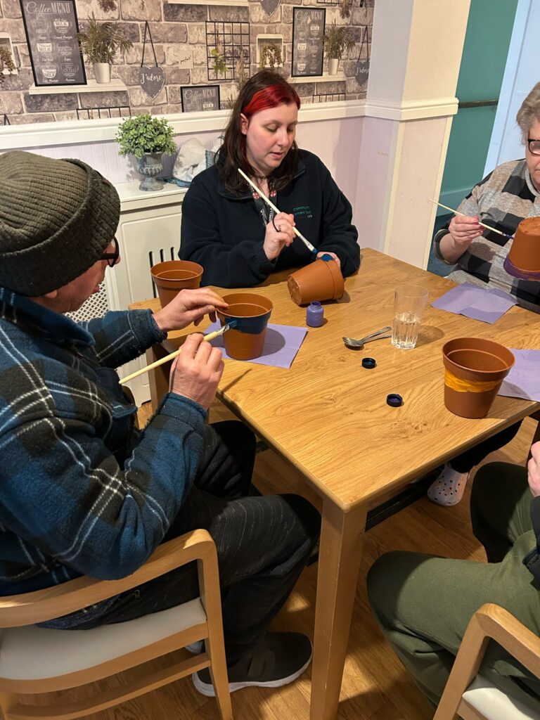 A close-up of three people painting terracotta plant pots at a wooden table. A woman with red and black hair is guiding the activity, while two other participants, one in a beanie and another in a striped top, focus on their designs. The table is covered with paint pots, brushes, and glasses of water.