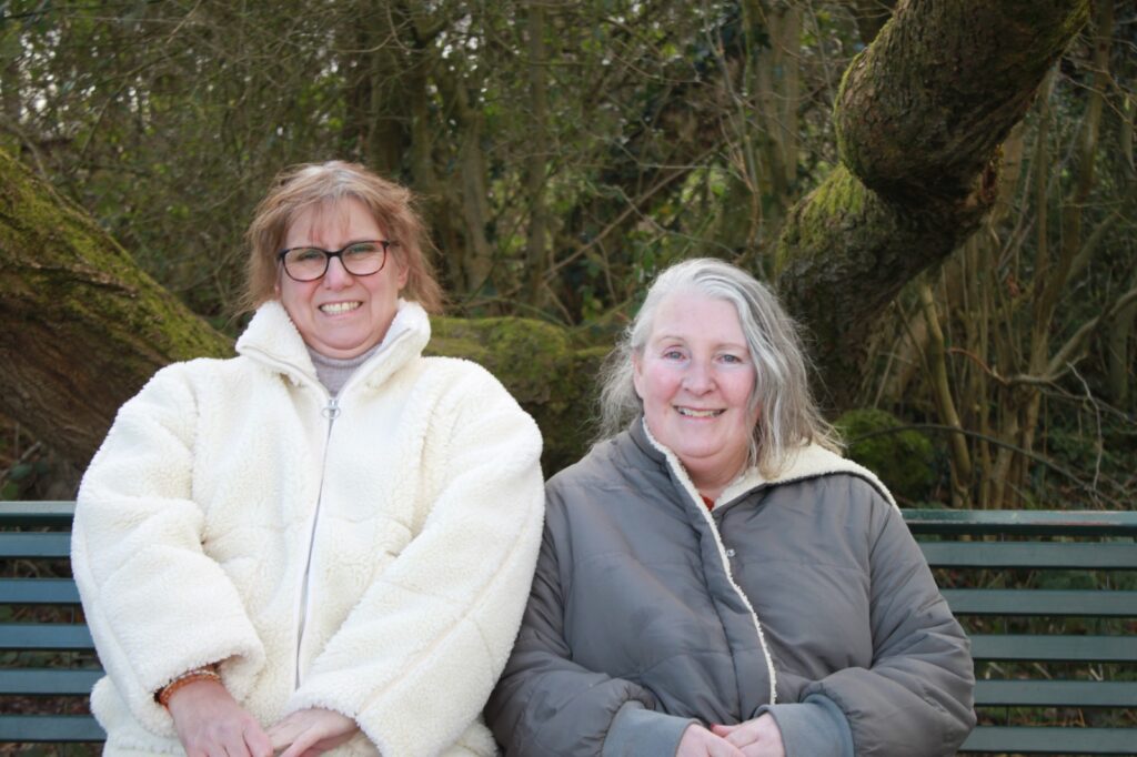 The two women sit side by side on a green park bench, leaning against a moss-covered tree trunk. Both smile warmly at the camera.