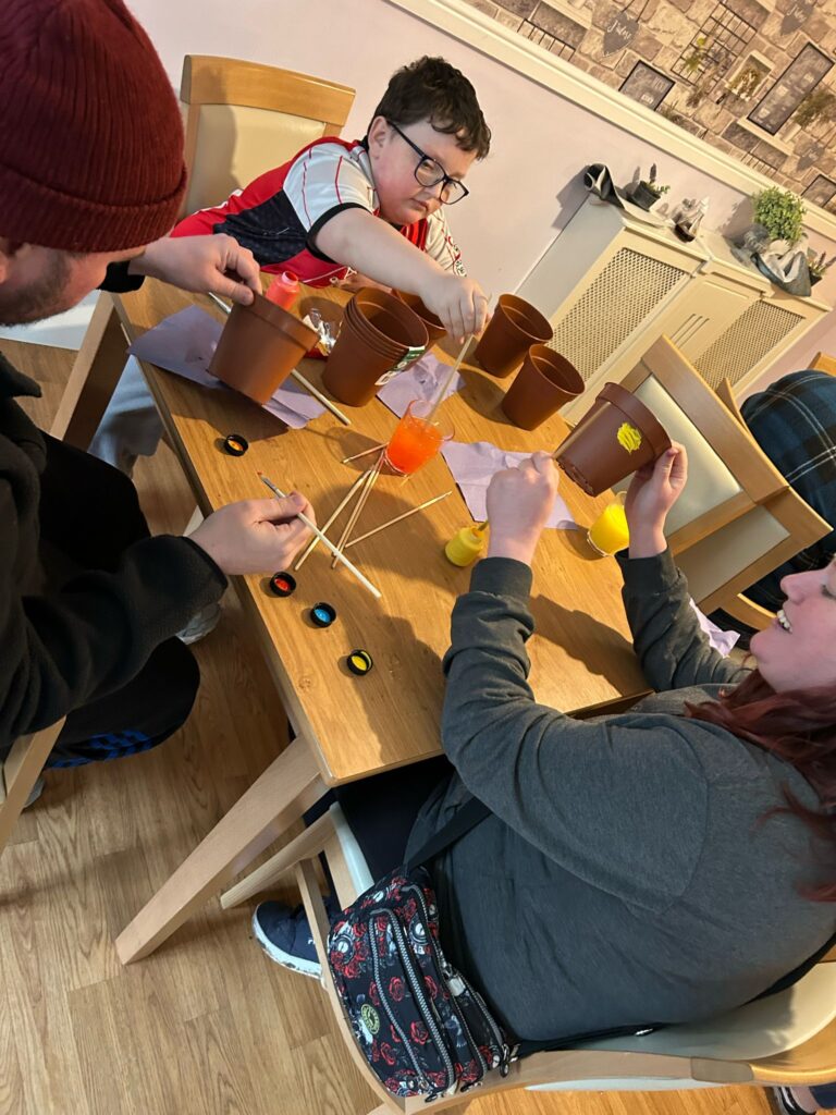 A young boy in glasses and a red football shirt is painting a terracotta pot with yellow paint, concentrating on his design. Two adults beside him are also painting pots, using brushes and different coloured paints. The table is scattered with small paint containers, napkins, and a glass of orange juice.